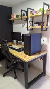 Escritorio de madera con marco metálico, acompañado de silla ergonómica y estanterías decorativas. Ideal para oficina en casa o setup de gaming, optimizando el espacio de trabajo y almacenamiento.