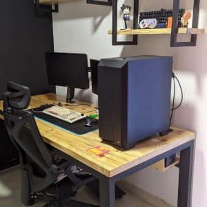 Escritorio de madera con marco metálico, acompañado de silla ergonómica y estanterías decorativas. Ideal para oficina en casa o setup de gaming, optimizando el espacio de trabajo y almacenamiento.