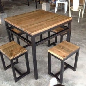 Mesa cuadrada con estructura metálica negra y superficie de madera, acompañada de cuatro taburetes, ideal para cocinas pequeñas o comedores contemporáneos.