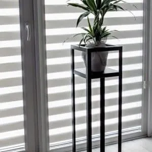 Soporte de metal negro minimalista con planta decorativa en maceta, ideal para embellecer espacios interiores modernos junto a ventanas grandes.