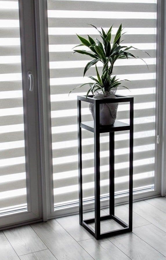 Soporte de metal negro minimalista con planta decorativa en maceta, ideal para embellecer espacios interiores modernos junto a ventanas grandes.