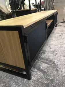 Mueble bajo de estilo industrial con estructura de metal negro y superficies de madera clara. Cuenta con puertas de rejilla y estantes abiertos para almacenamiento.