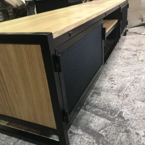 Mueble bajo de estilo industrial con estructura de metal negro y superficies de madera clara. Cuenta con puertas de rejilla y estantes abiertos para almacenamiento.