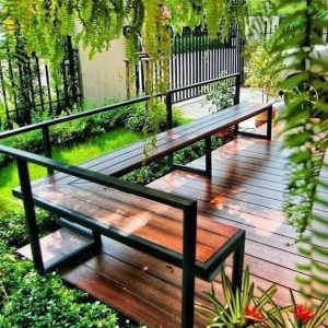 Banco de jardín moderno con estructura metálica negra y asiento de madera sobre terraza, rodeado de vegetación exuberante. Diseño minimalista y elegante para exteriores.