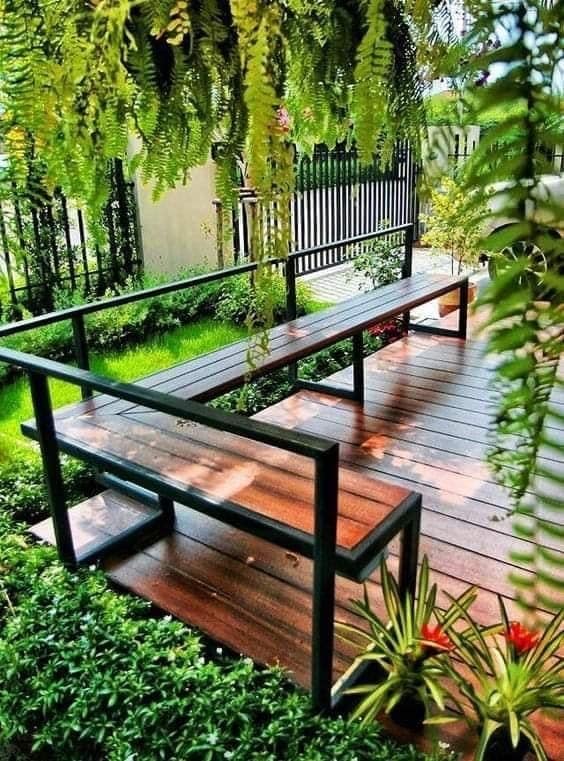 Banco de jardín moderno con estructura metálica negra y asiento de madera sobre terraza, rodeado de vegetación exuberante. Diseño minimalista y elegante para exteriores.