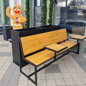 Banco de exterior moderno con estructura metálica negra y asientos de madera clara. Incluye mesas pequeñas integradas en el diseño, ideal para áreas recreativas urbanas.