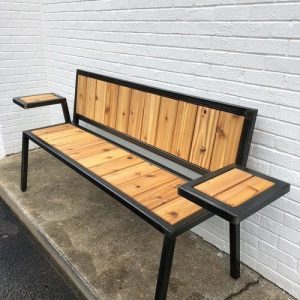 Banco moderno con estructura de metal negro y superficie de madera clara. Cuenta con respaldo y dos superficies laterales de apoyo. Ideal para exteriores o jardines.