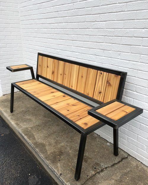 Banco moderno con estructura de metal negro y superficie de madera clara. Cuenta con respaldo y dos superficies laterales de apoyo. Ideal para exteriores o jardines.