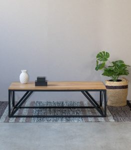 Mesa larga con superficie de madera y patas de metal negro en diseño geométrico. Colocada sobre alfombra, decorada con una piña cerámica y libros. Decorativa y moderna.