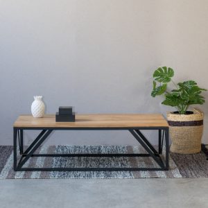 Mesa larga con superficie de madera y patas de metal negro en diseño geométrico. Colocada sobre alfombra, decorada con una piña cerámica y libros. Decorativa y moderna.