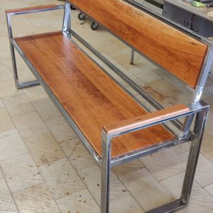 Banco de diseño industrial, con estructura de metal pulido y asiento de madera. Ideal para interiores modernos, combina funcionalidad y estilo contemporáneo.