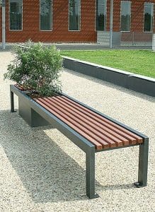 Banco de exterior con estructura de metal negro y listones de madera. Incluye una maceta integrada con arbusto. Diseño moderno y minimalista, ubicado en un área pavimentada.