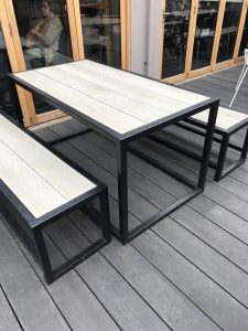 Mesa de exterior moderna con estructura metálica negra y tablero de madera clara, acompañada de bancos a juego, ubicada sobre un piso de madera gris.