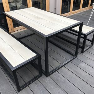 Mesa de exterior moderna con estructura metálica negra y tablero de madera clara, acompañada de bancos a juego, ubicada sobre un piso de madera gris.