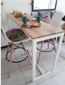 Mesa alta de cocina con superficie de madera clara y estructura blanca metálica, acompañada de sillas altas de patas de madera y asiento blanco, ideal para desayunos.