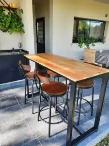 Mesa alta de madera con estructura metálica negra acompañada de cuatro sillas altas con respaldo de madera y patas metálicas, ubicada en un espacio moderno de terraza.