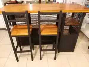 Mesa alta de madera con dos sillas a juego. La estructura es metálica con estantes laterales y un diseño de rejilla negra. Adecuada para cocina o bar.