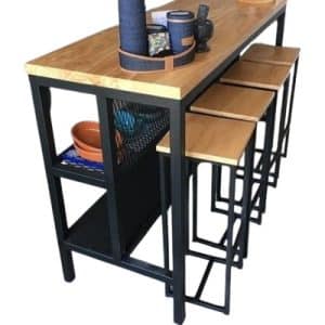 Mesa alta de madera clara con estructura metálica negra, incluye cuatro taburetes a juego, ideal para cocina o bar. Diseño moderno con un estante lateral de rejilla.