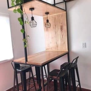 Mesa alta con estructura de metal negro y superficie de madera clara. Incluye dos lámparas colgantes y estantes superiores para plantas. Acompañada de taburetes negros. Ideal para cocinas.