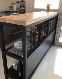 Una isla de cocina moderna con encimera de madera clara y estructura metálica negra, incluye estantes de rejilla para almacenamiento de botellas y platos.