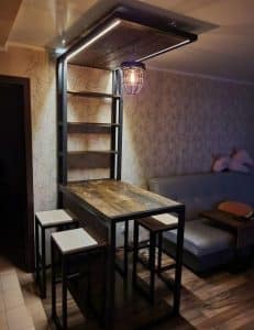 Mesa alta de madera con estructura metálica, acompañada de taburetes y estantes en conjunto; diseño industrial con iluminación colgante, ideal para espacios modernos y funcionales.