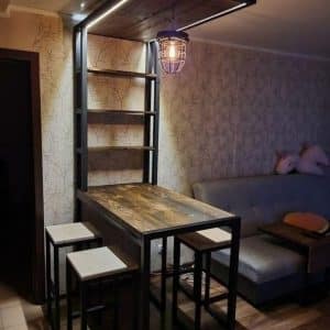 Mesa alta de madera con estructura metálica, acompañada de taburetes y estantes en conjunto; diseño industrial con iluminación colgante, ideal para espacios modernos y funcionales.