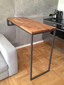 Mesa alta de madera con soporte lateral de metal negro, diseño minimalista flotante, sobre un suelo de parquet claro.