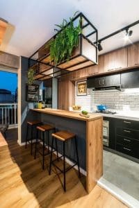 Barra de cocina moderna de madera con tres taburetes altos. Diseño minimalista con estantería suspendida de metal negro y decoración de plantas colgantes.