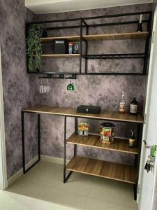 Mueble de bar esquinero con estructura de metal negro y estantes de madera, incluye estantes para copas y botellas en una pared con acabado gris texturizado.