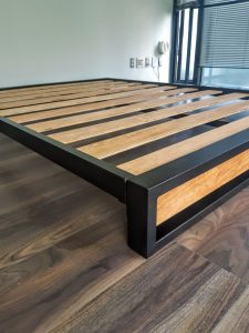 Estructura de cama moderna con marco de metal negro y listones de madera, sobre piso de madera oscura. Diseño minimalista y funcional, ideal para un dormitorio contemporáneo.