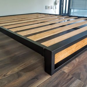 Estructura de cama moderna con marco de metal negro y listones de madera, sobre piso de madera oscura. Diseño minimalista y funcional, ideal para un dormitorio contemporáneo.