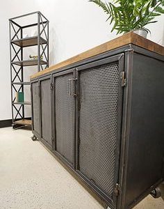 Gabinete industrial de metal con cuatro puertas de rejilla y superficie de madera. Está montado sobre ruedas para facilitar su movilidad. Ideal para espacios modernos y funcionales.
