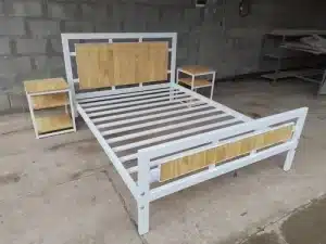 Cama de estructura metálica blanca con detalles de madera clara en el cabecero y pie de cama, acompañada de dos mesitas de noche a juego en un entorno industrial.