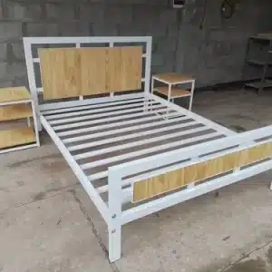 Cama de estructura metálica blanca con detalles de madera clara en el cabecero y pie de cama, acompañada de dos mesitas de noche a juego en un entorno industrial.