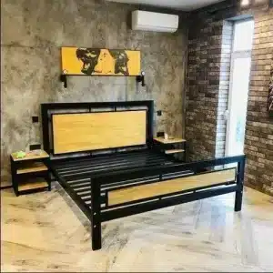 Cama de metal y madera con cabecero y pieceros rectangulares. Incluye mesas de noche a juego y se sitúa en una habitación con paredes de ladrillo y suelo de madera.