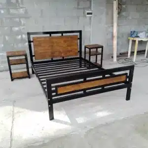 Cama de metal con detalles de madera en el cabecero y pie, acompañada de dos mesas de noche a juego, situada en un entorno industrial con paredes de bloques grises.