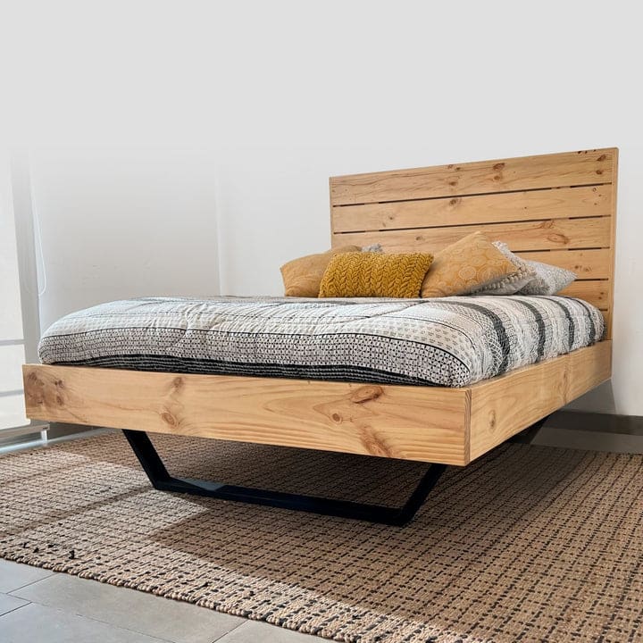 Cama moderna de madera clara con cabecera de tablones horizontales. Base flotante sobre patas negras angulares. Colchón cubierto por colcha y cojines en tonos beige y amarillo.