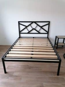 Estructura de cama de metal negro con cabecera geométrica y listones de madera, situada en una habitación con suelo laminado.