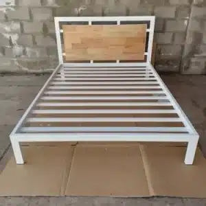 Estructura de cama individual moderna con marco de metal blanco y cabecero de madera clara sobre piso de concreto.