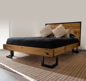 Cama moderna de madera natural con cabecera rústica. Base robusta de metal negro y colchón oscuro, complementada con cojines texturizados. Ideal para un dormitorio contemporáneo.