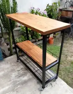 Carrito de cocina con estantes de madera y estructura metálica negra. Tiene ruedas para fácil movilidad y ofrece dos niveles de almacenamiento. Ideal para uso interior o exterior.