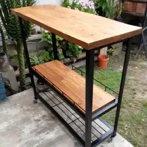 Carrito de cocina con estantes de madera y estructura metálica negra. Tiene ruedas para fácil movilidad y ofrece dos niveles de almacenamiento. Ideal para uso interior o exterior.
