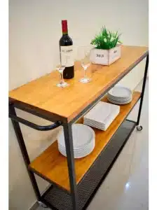 Carrito de cocina con estructura metálica negra y tres estantes de madera. Incluye platos, copas y una caja decorativa sobre la superficie superior.