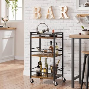 Carrito de bar de metal y madera de tres niveles con ruedas, decorado con botellas y copas, ideal para espacios de cocina o sala, junto a una pared de ladrillo pintada de blanco.