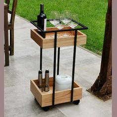 Carrito de servicio de madera con estructura metálica negra, dos niveles, superior con copas y botella, inferior con salero y pimentero. Ideal para exteriores o eventos.