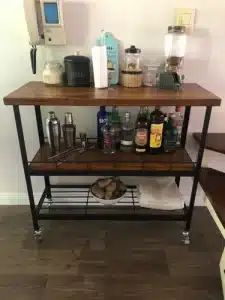 Carrito de bar de dos niveles con estructura de metal negro y superficies de madera, equipado con ruedas, contiene botellas, tarros y utensilios de coctelería. Ideal para organizar bebidas.