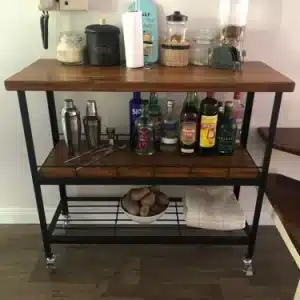 Carrito de bar de dos niveles con estructura de metal negro y superficies de madera, equipado con ruedas, contiene botellas, tarros y utensilios de coctelería. Ideal para organizar bebidas.