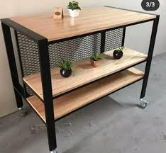 Mesa industrial con estructura de metal negro y superficie de madera, dos estantes inferiores, ruedas para movilidad, ideal para decoración moderna o uso en cocina o taller.