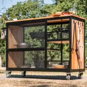 Carrito de cocina de exterior en madera y metal, con ruedas. Incluye superficie de trabajo, estantes y ganchos laterales. Ideal para asados o uso en jardín.