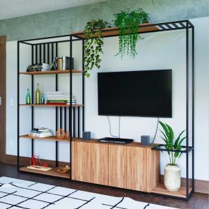 Unidad de entretenimiento moderna con estanterías de madera y metal negro. Incluye espacio para TV, decorada con plantas y libros en los estantes. Diseño abierto y estilizado.